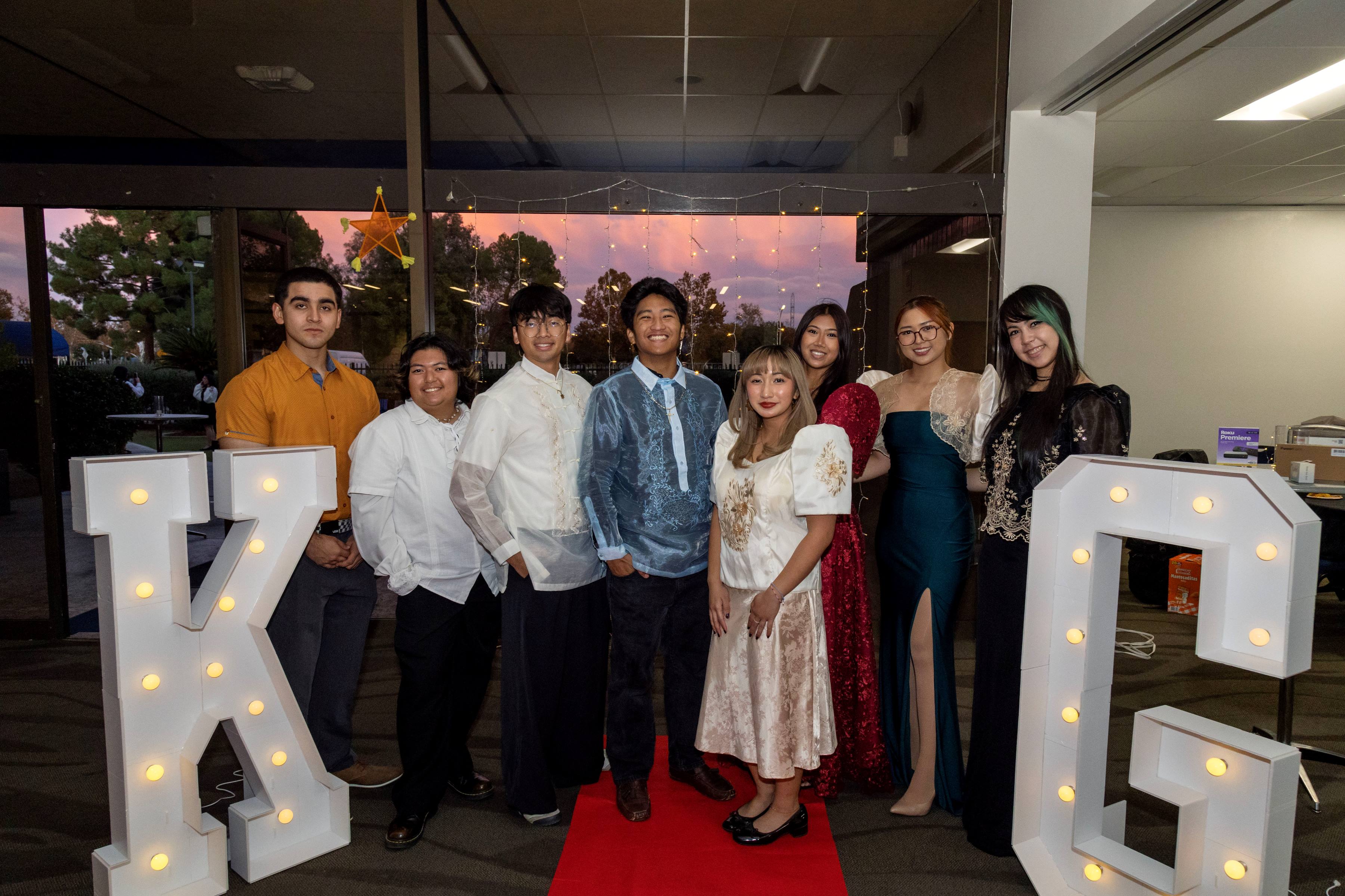 Group of people in between in front of big white letters with lightbulbs in them stating "K" and "G". There is a red carpet rolled between the K and G