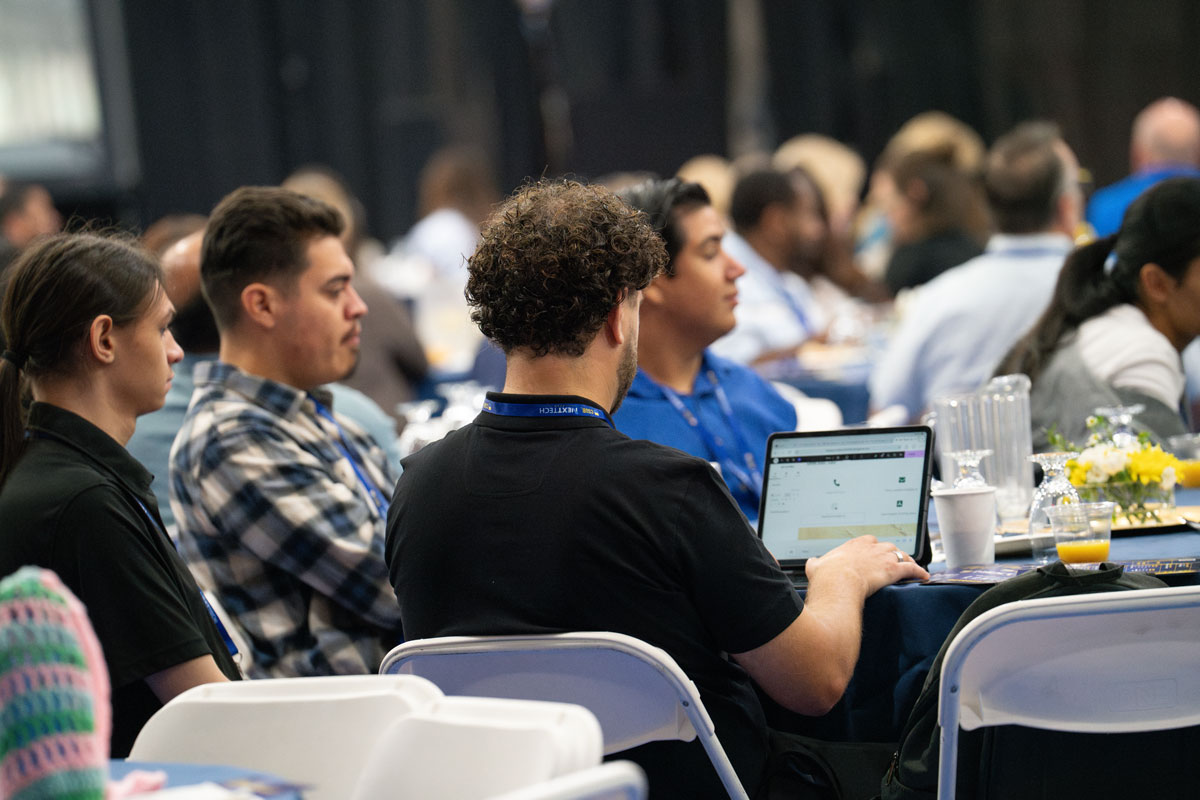 A group of attendees sit at a table during a conference, one person working on a laptop.