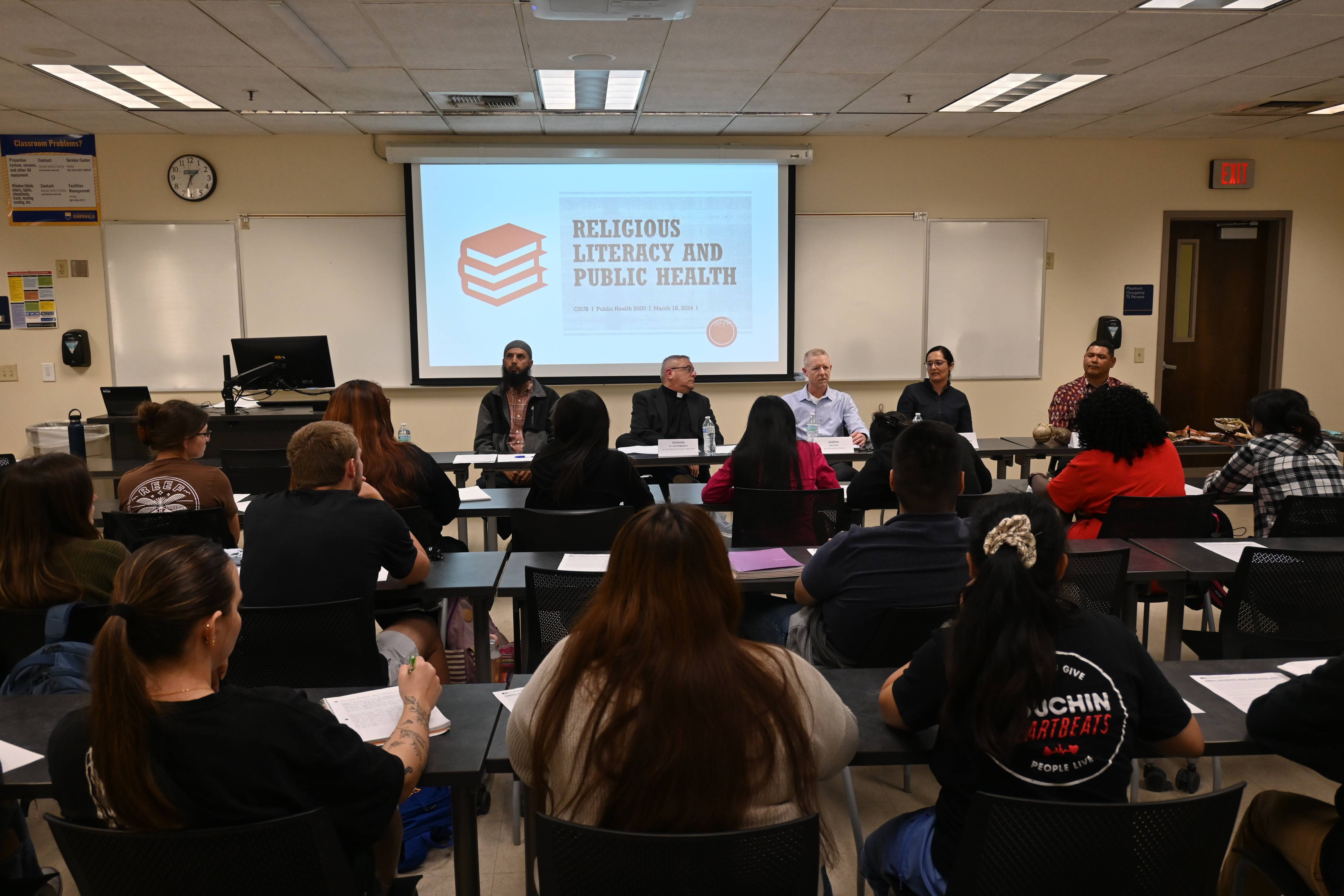 Religious leaders speak at the front of the classroom. Students sit at tables listening.